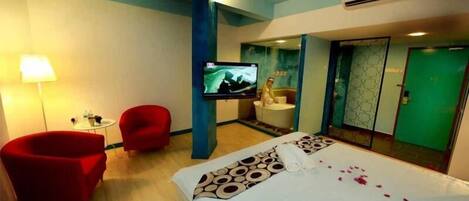 A hotel room with a bed, a TV, a red armchair, a small table, and a bathroom visible through an open door.