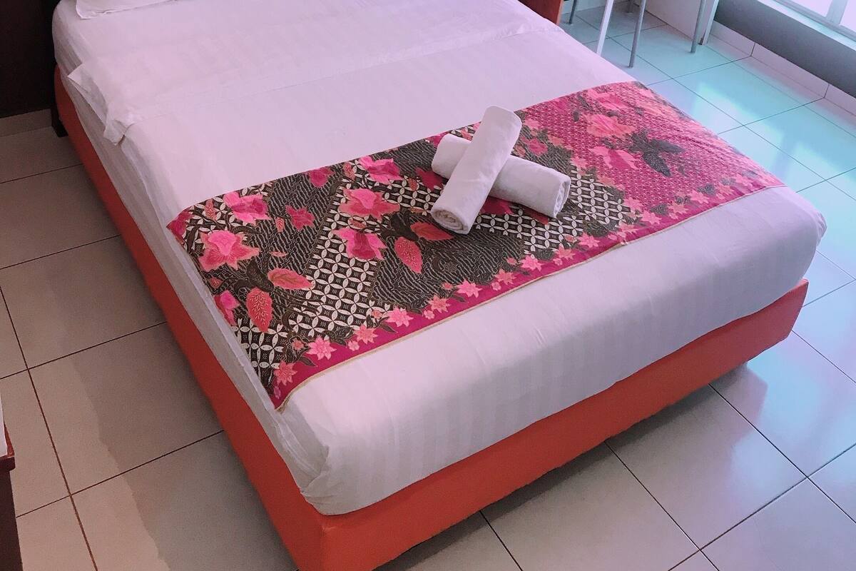 A neatly made bed with white linens and a floral-patterned bedspread, a small desk with a chair, a telephone, and a bottle of water on the desk.