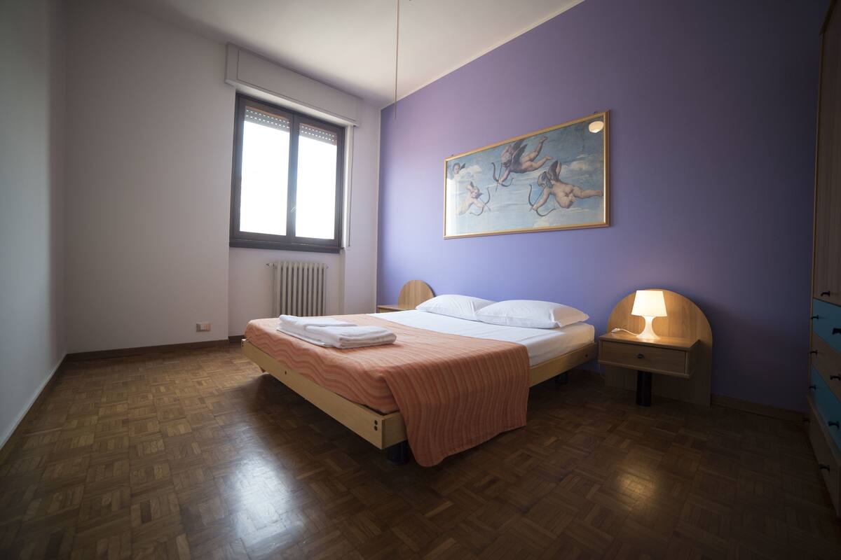 A bedroom with a wooden floor, a bed with a pink bedspread, a nightstand with a lamp, a window with blinds, and a painting on the wall.