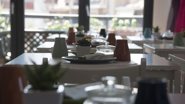 A dining area with tables set for two, featuring white tablecloths, plates, and cutlery. There are potted plants and a view of buildings through large windows. - Alex e Angie (Bergamo)