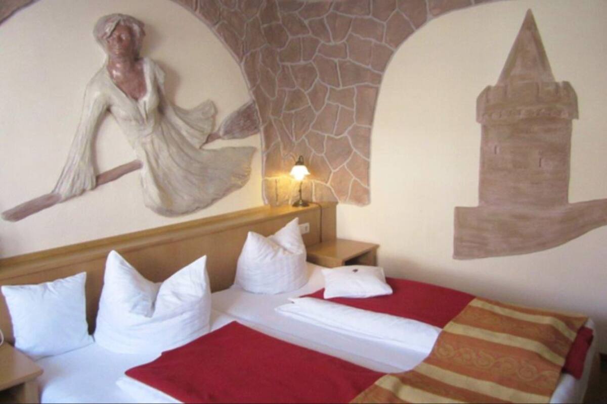Ein Hotelzimmer mit Bett, Kissen, einer roten Decke, einem Nachttisch, einer Wandleuchte und einer Steinwand mit Wandgemälde.