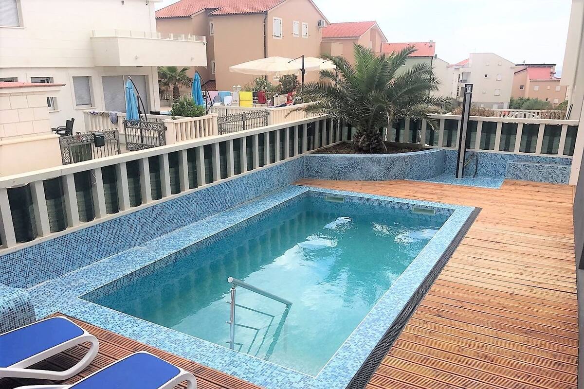 A rooftop pool with blue-tiled edges, surrounded by a wooden deck and blue lounge chairs.