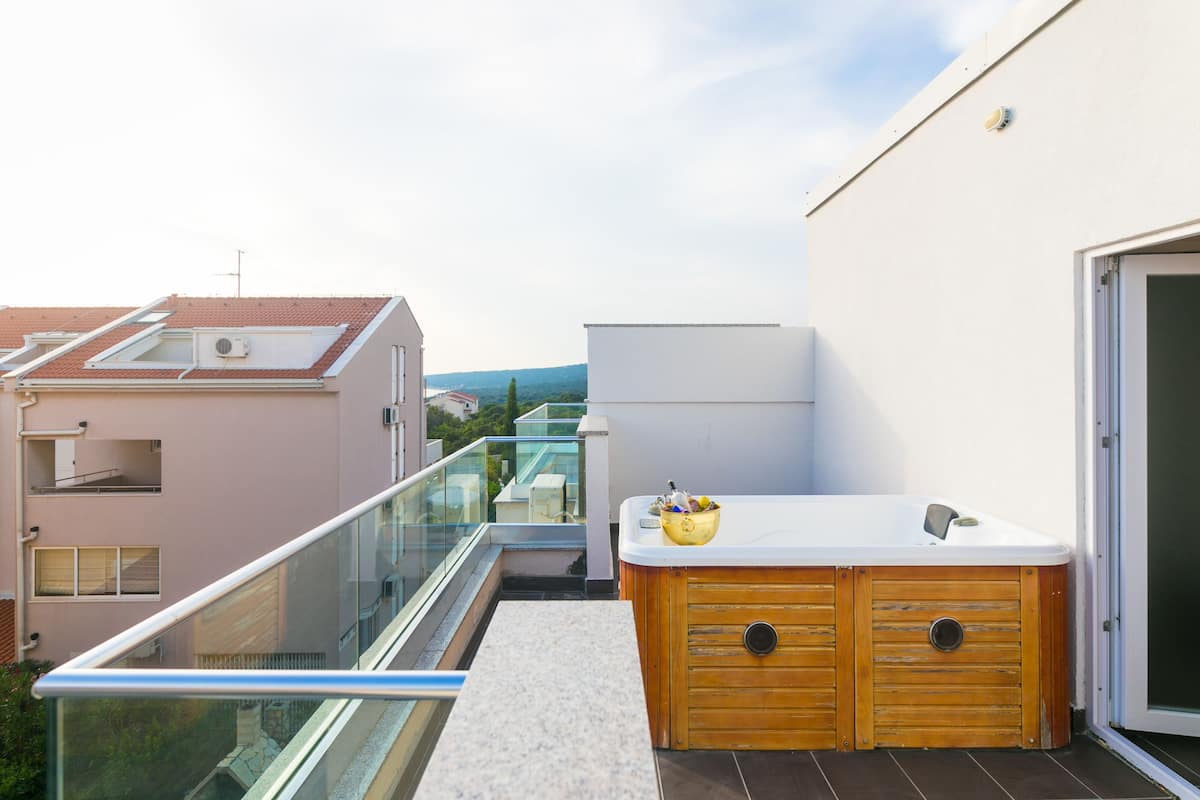 A rooftop terrace with a hot tub, glass balustrade, and a view of neighboring buildings.