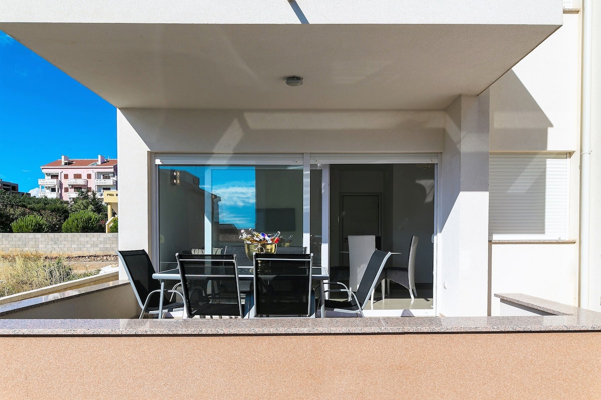 A modern outdoor balcony with a glass door, a table with chairs, and a decorative bowl.
