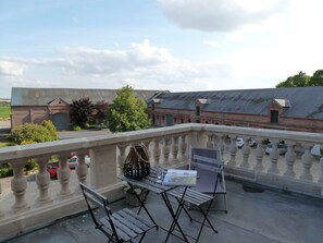A balcony with a table and chairs, a metal fence, and a view of buildings and parked cars. - Ferme de Bonavis - Chambres d'Hôtes (Banteux)