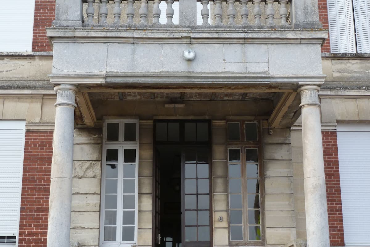 A building with a stone facade, a central entrance with a dark door, and two columns flanking the entrance.