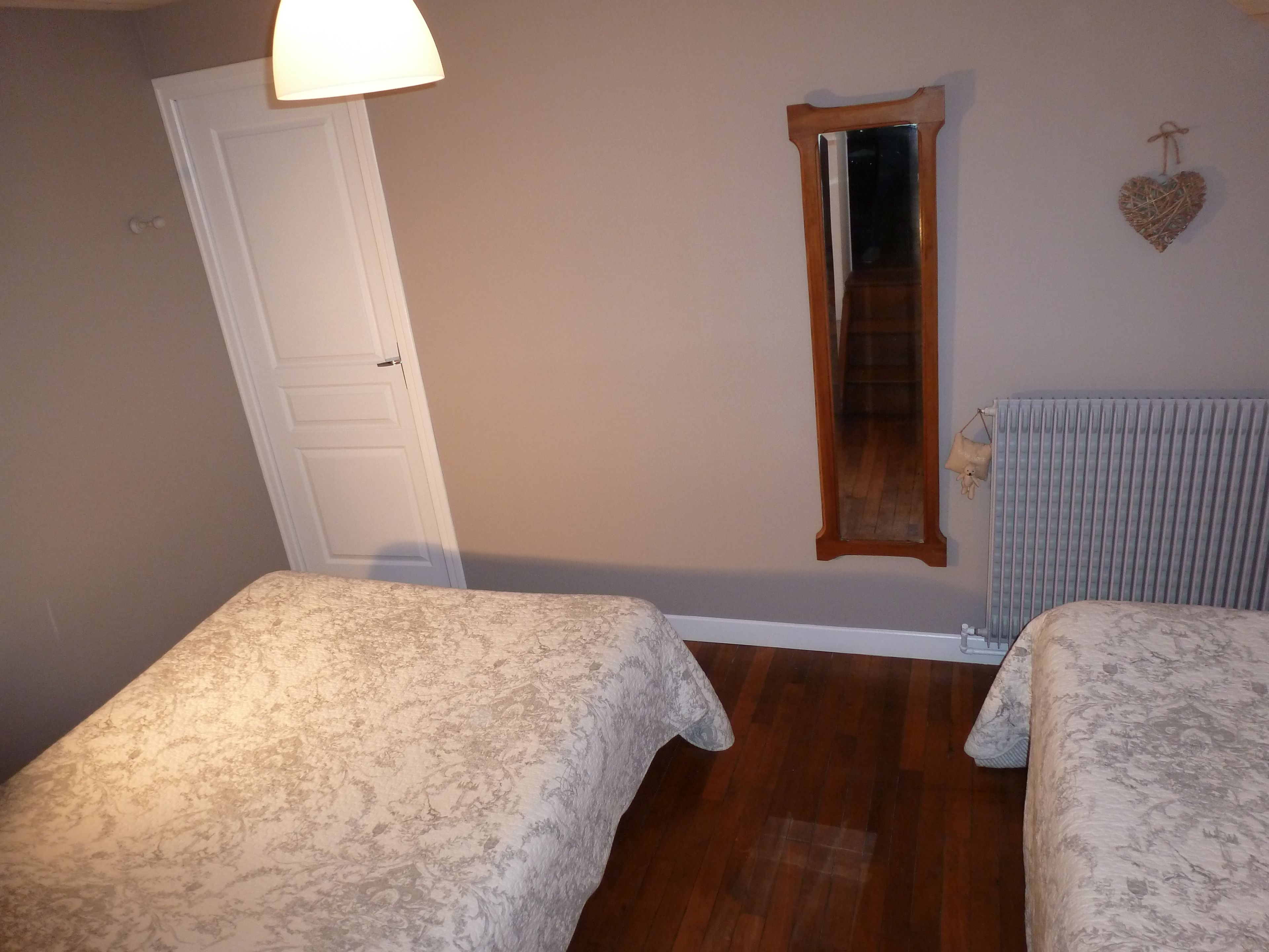 A room with two beds, a door, a mirror, a radiator, and a heart-shaped decoration on the wall.