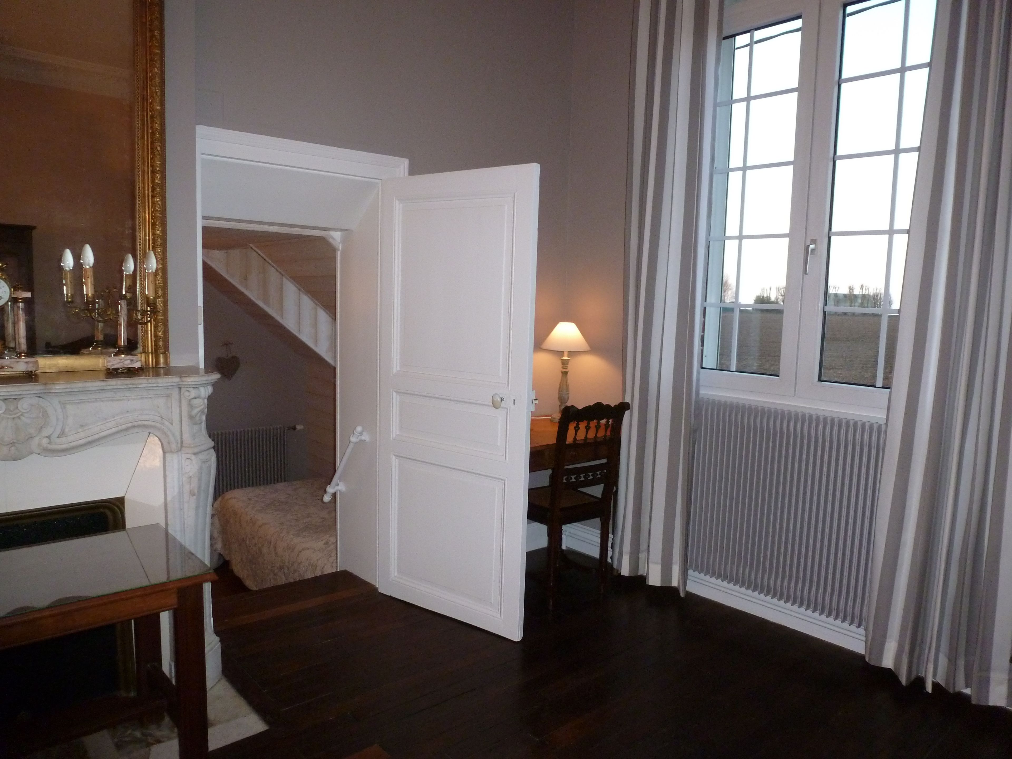 A room with a wooden floor, a white door, a table with a lamp, and a window with curtains.