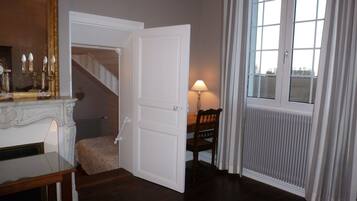 A room with a wooden floor, a white door, a table with a lamp, and a window with curtains.