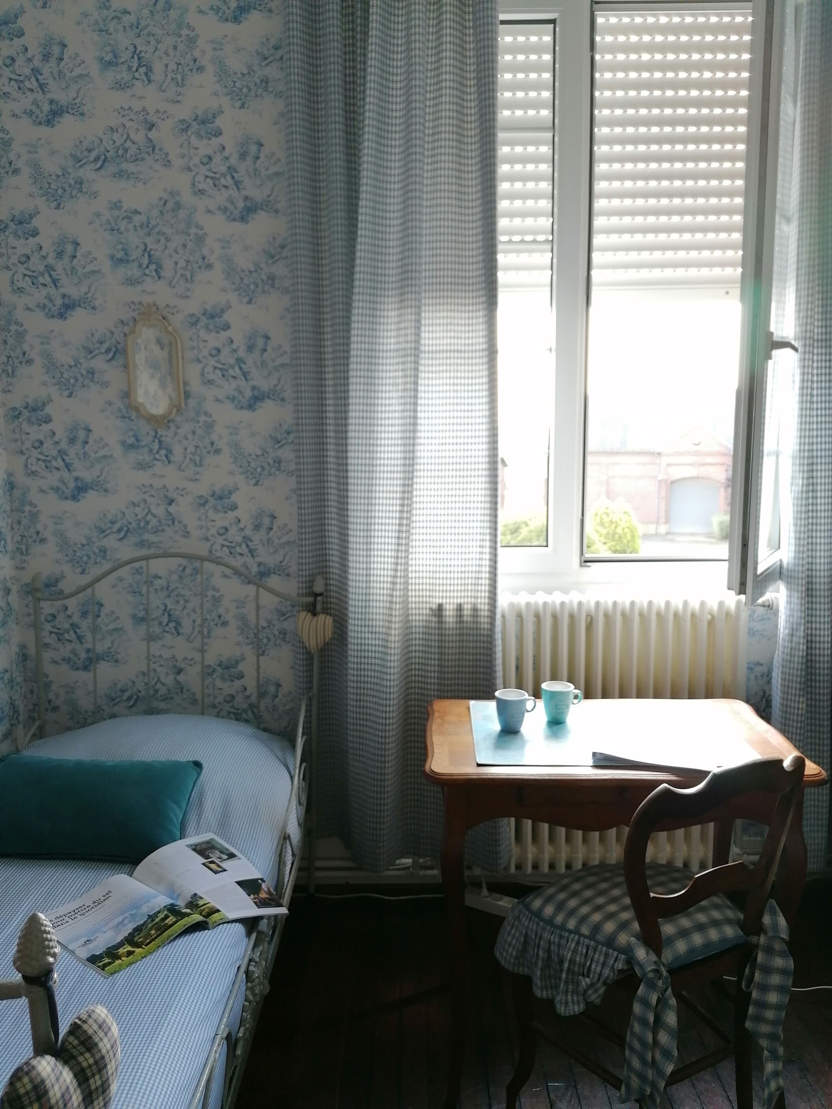 A bedroom with a bed, a wooden table with two cups, a chair, a radiator, and a window with blinds.