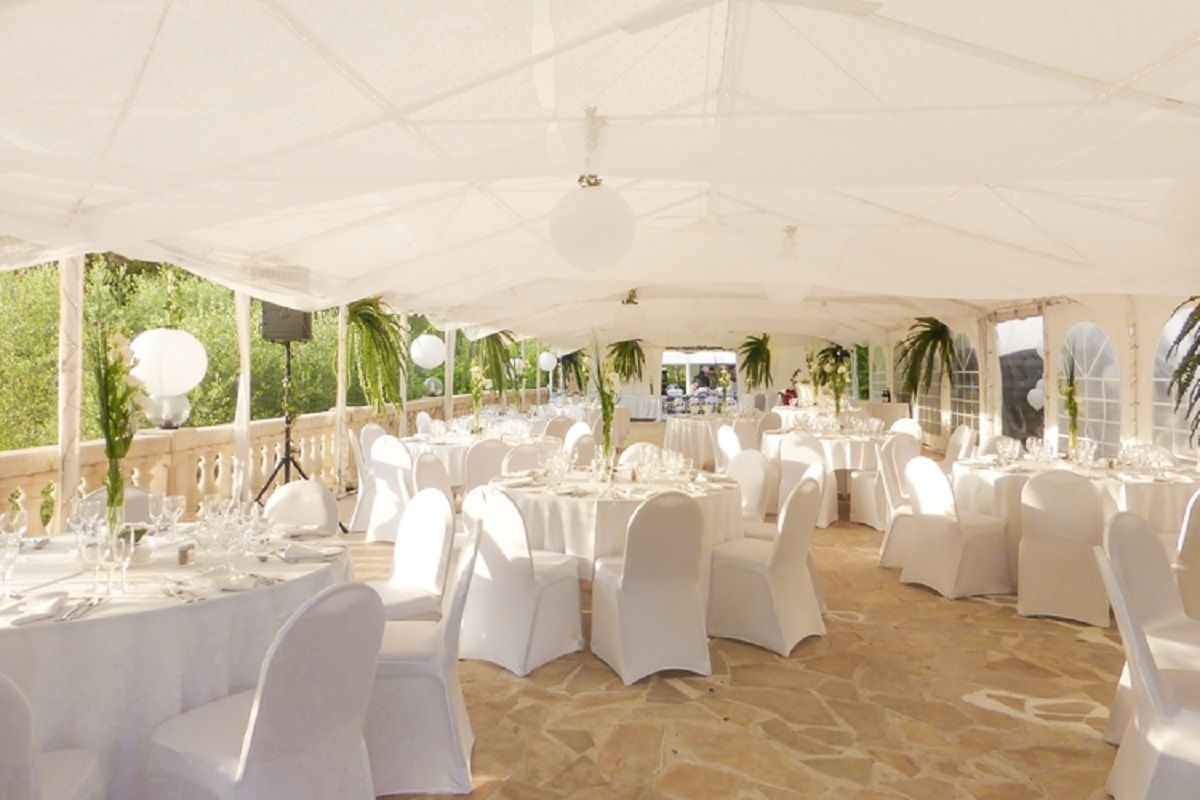 An outdoor banquet hall with round tables set for dining, covered by a white canopy, and surrounded by palm trees.