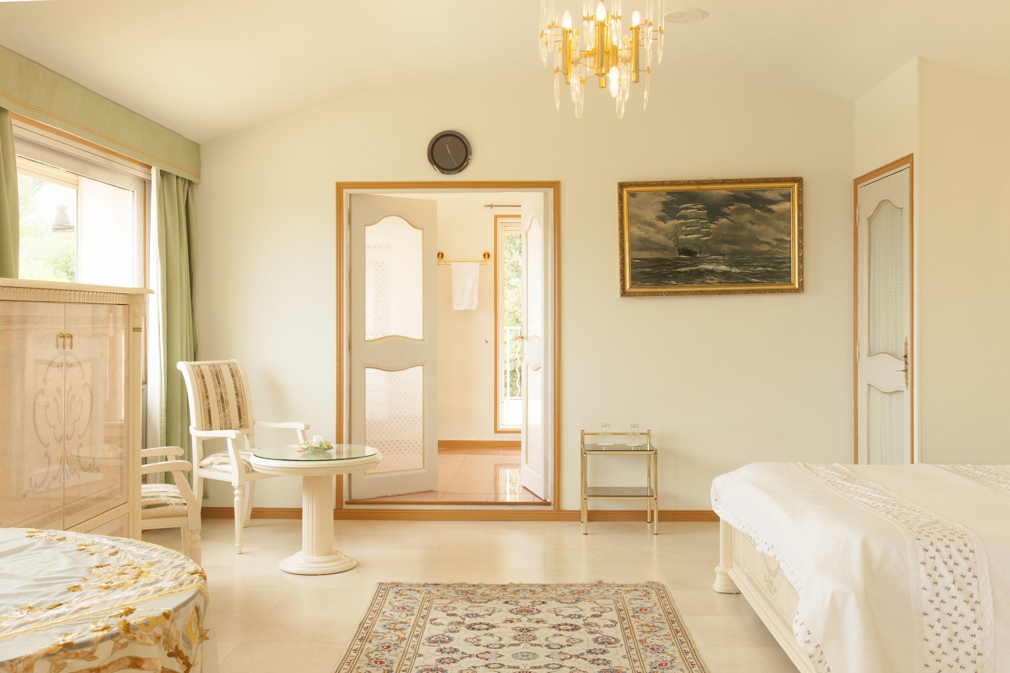 A hotel room with a bed, a chair, a small table, a chandelier, a painting, and a view of the outside.