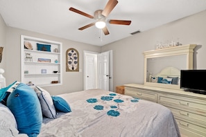 Une chambre à coucher comprenant un lit, un ventilateur de plafond, une commode, un téléviseur et une bibliothèque.