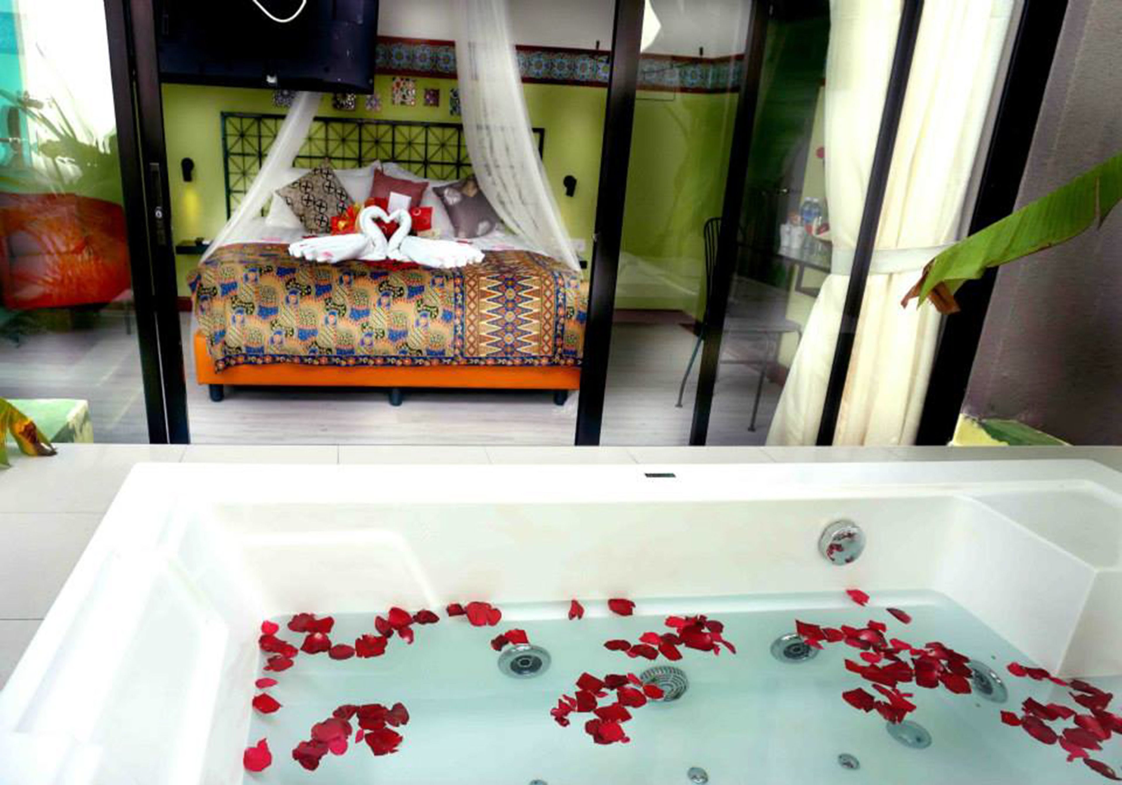 A bedroom with a bed covered in a colorful patterned bedspread, a canopy, and a heart-shaped decoration. In front of the bed, there is a bathtub filled with water and rose petals.