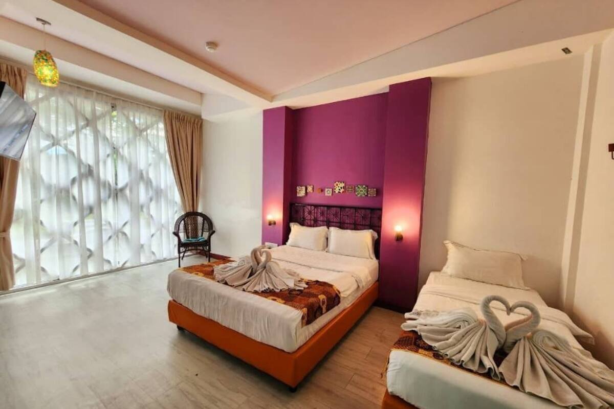Quarto de hotel com duas camas, uma janela grande com cortinas, uma cadeira e uma parede de destaque roxa.