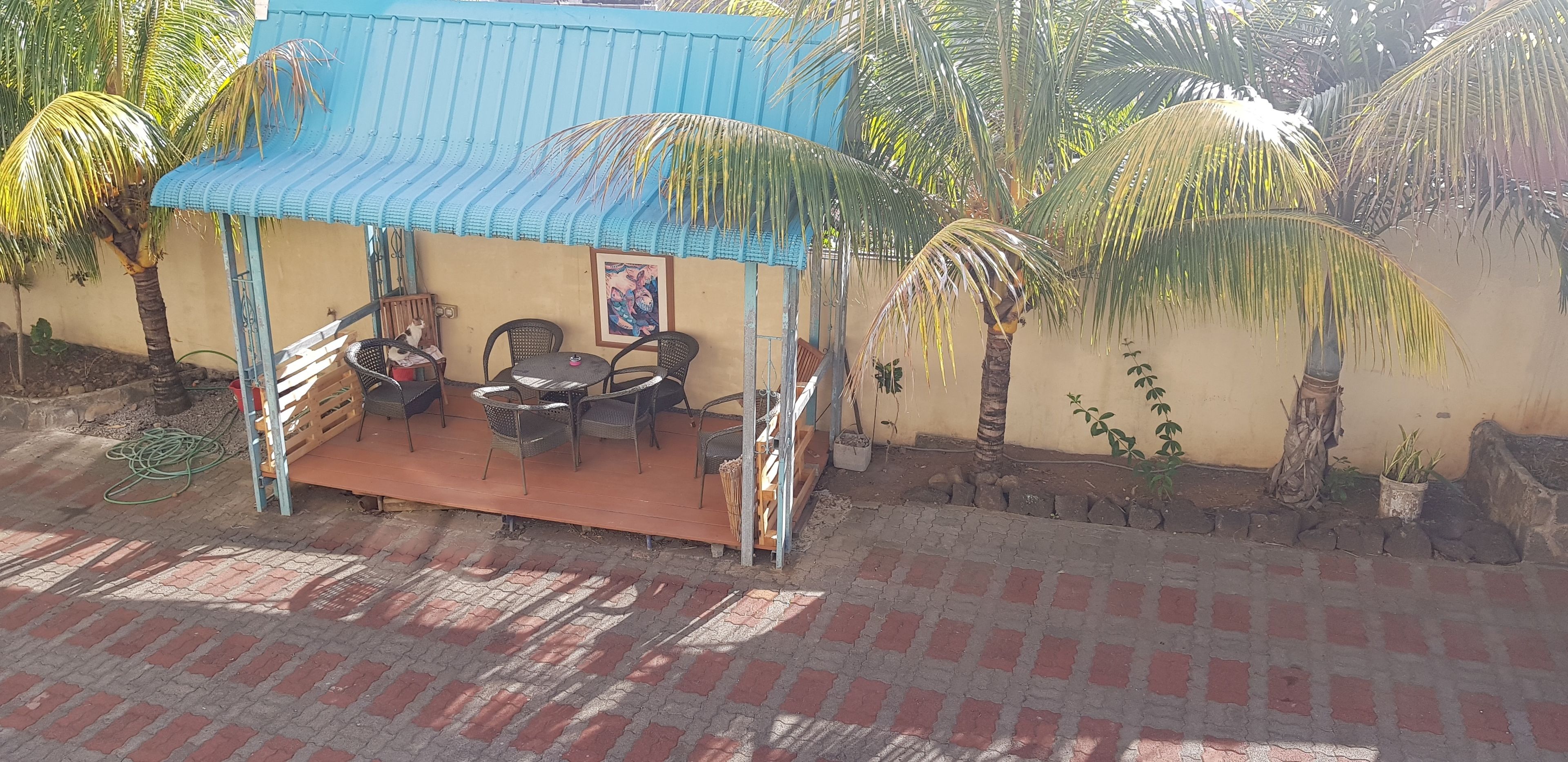 Un espacio al aire libre techado con sillas y una mesa, rodeado de palmeras y una estructura con techo azul.
