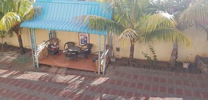 Un espacio al aire libre techado con sillas y una mesa, rodeado de palmeras y una estructura con techo azul.