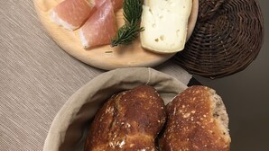 A wooden board with sliced ham, cheese, and a sprig of rosemary. Below, there is a basket containing two loaves of bread. - Atmosphere di Montagna (Vermiglio)