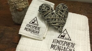 Two white towels with "Atmosphere Montagna" printed on them, a small decorative tree, and a woven heart-shaped object. - Atmosphere di Montagna (Vermiglio)