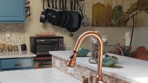 Una cocina con grifo de cobre, pileta negra y encimera de madera. Hay alacenas, horno tostador y un colgador para ollas.