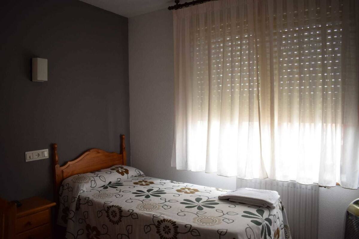Une chambre à coucher avec un lit en bois, une table de chevet, une fenêtre avec des rideaux légers et une lampe fixée au mur.