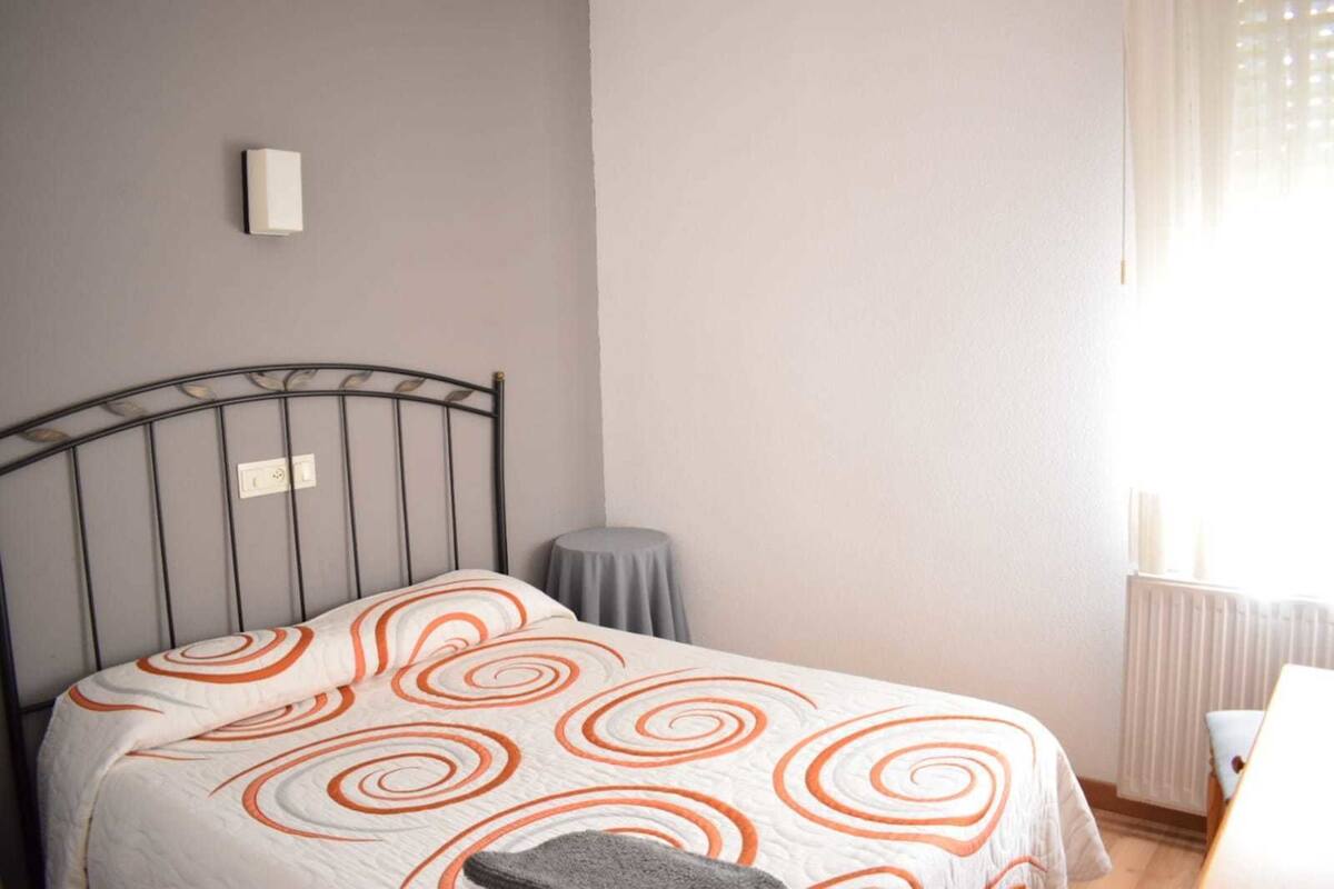 Une chambre à coucher avec un lit, une petite table, un radiateur et une fenêtre avec des rideaux.