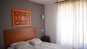 A bedroom with a wooden headboard, a bed with patterned bedding, a wall-mounted artwork, a bedside table, and a window with sheer curtains. - Hostal La Tablada (Navaleno)