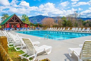 A swimming pool with lounge chairs and umbrellas, a wooden building with a green roof, and mountains in the background. - HEAVENLY DAZE — THREE KING SUITES, THREE LIVING AREAS & ROOM FOR 12 (Sevierville)