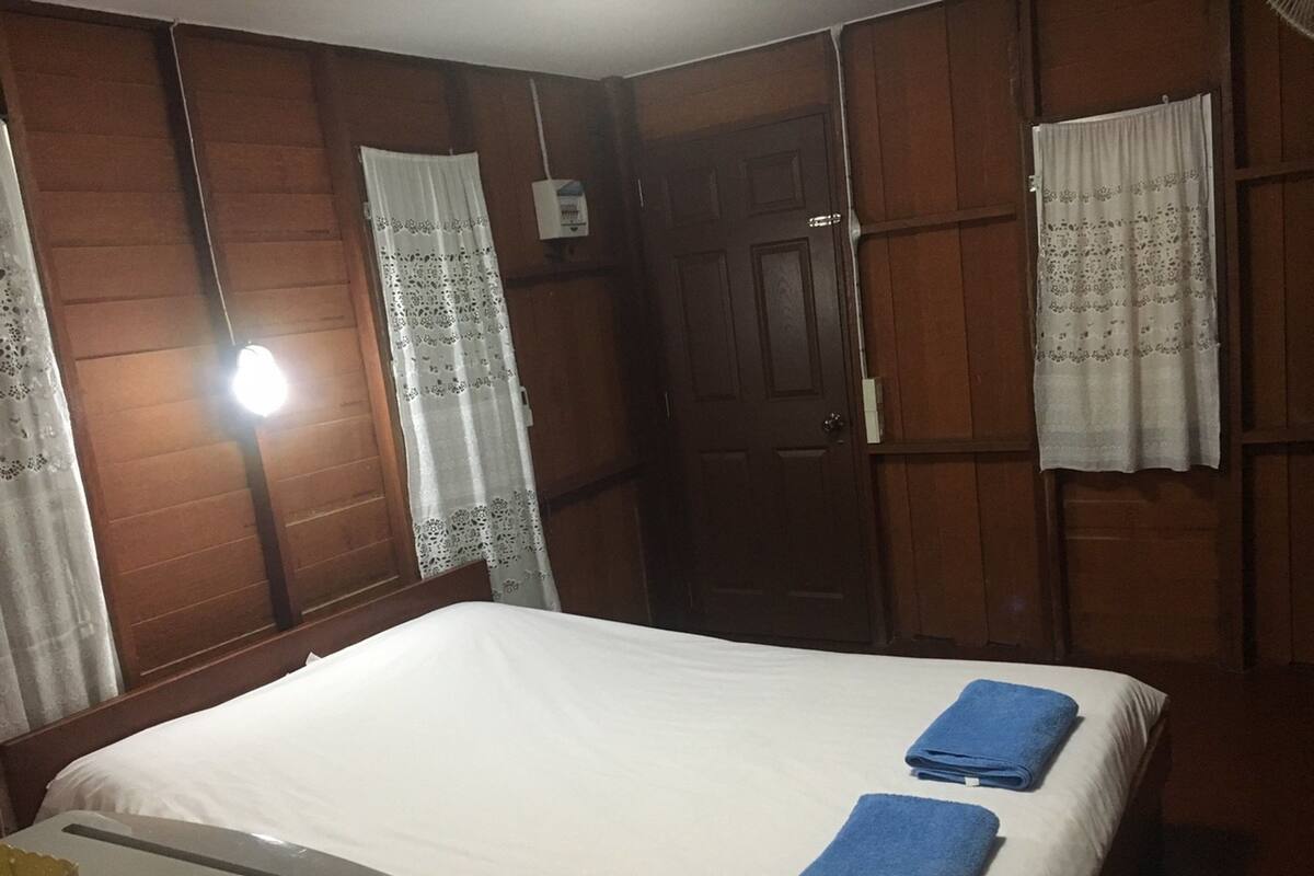 A hotel room with a bed, two blue towels, wooden panel walls, a door, and a window with a curtain.