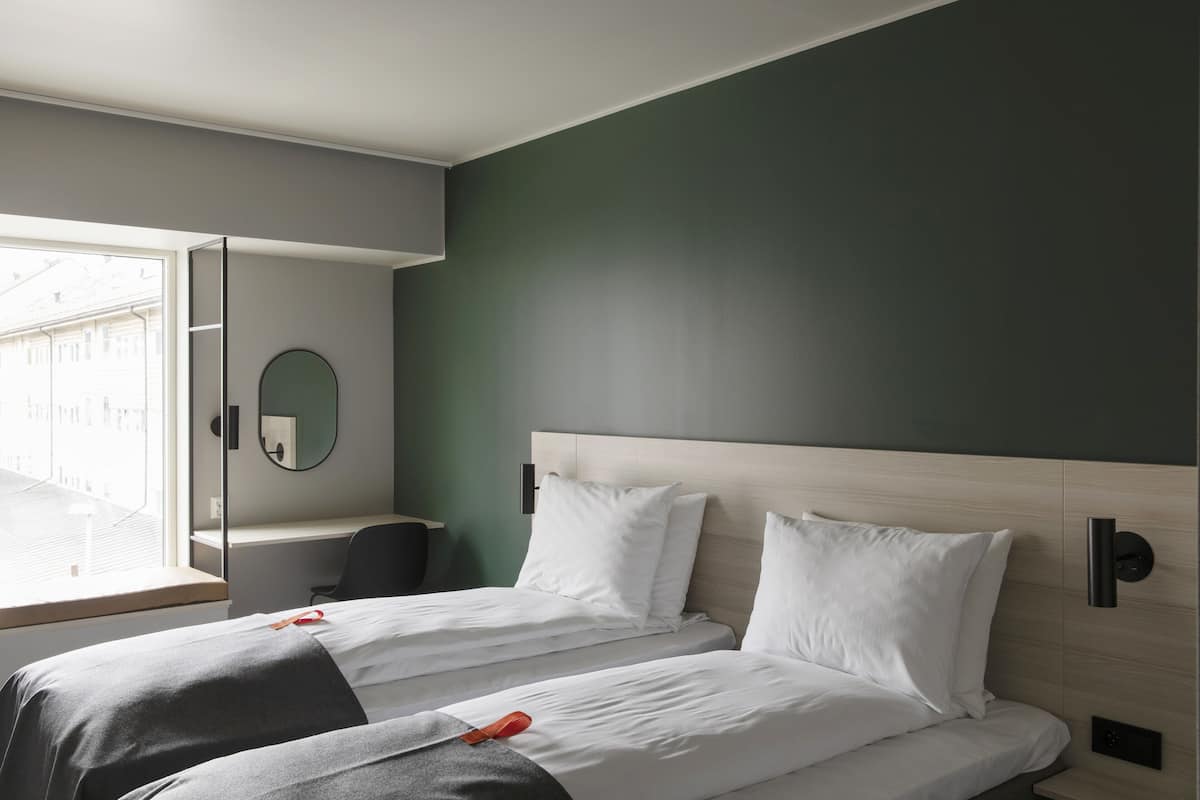Habitación de hotel moderna con dos camas, un escritorio, un espejo y una ventana con vistas a edificios.