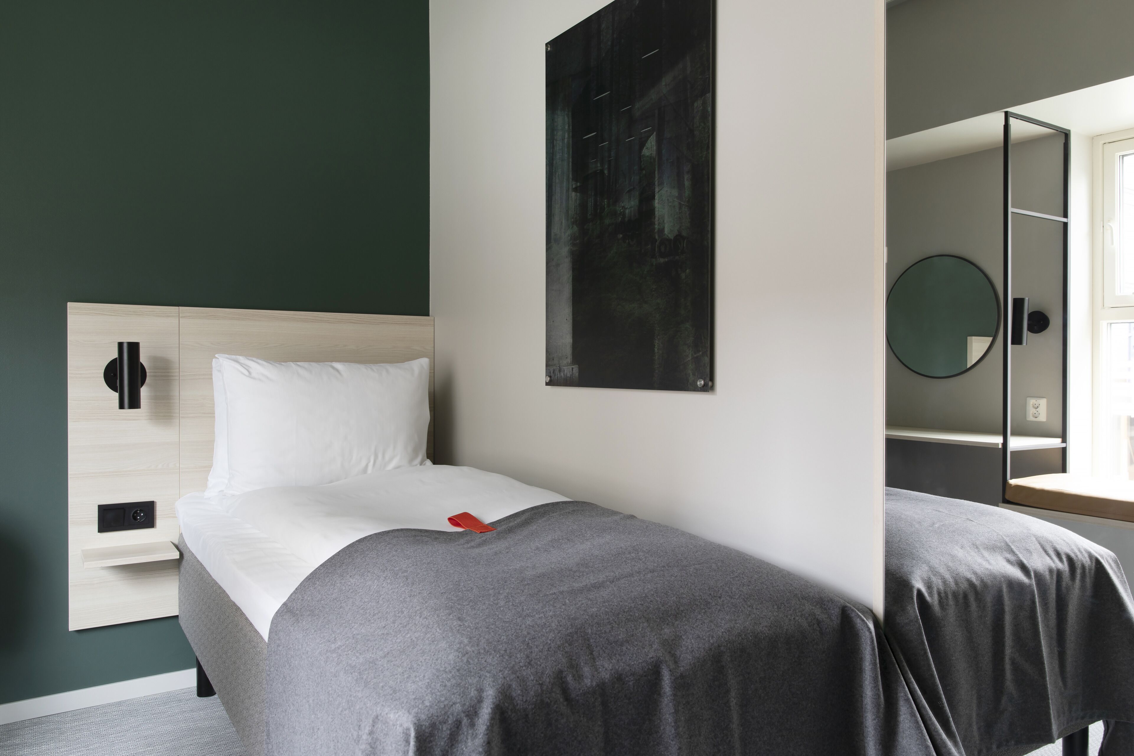 A hotel room with a bed, a bedside table, a mirror, and a wall-mounted artwork.