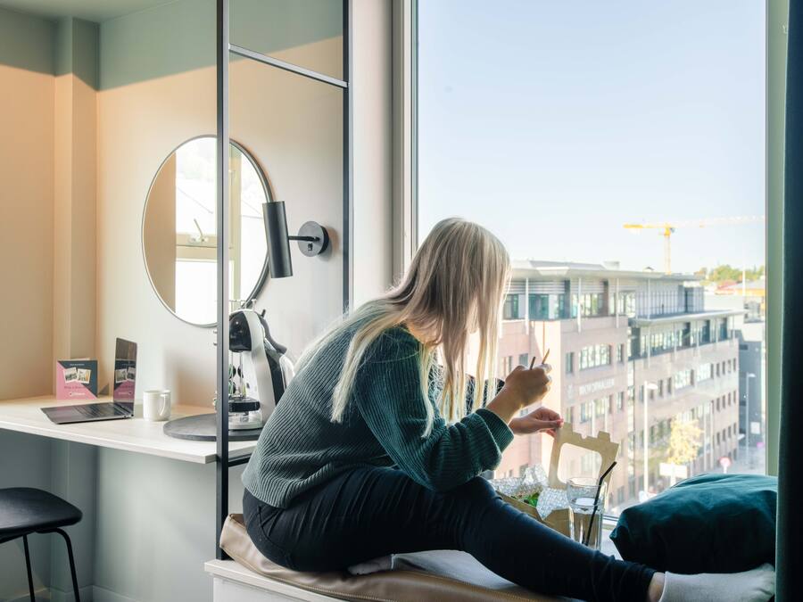 Una persona sentada junto a una ventana con una computadora portátil, una taza y un cuaderno sobre un escritorio.