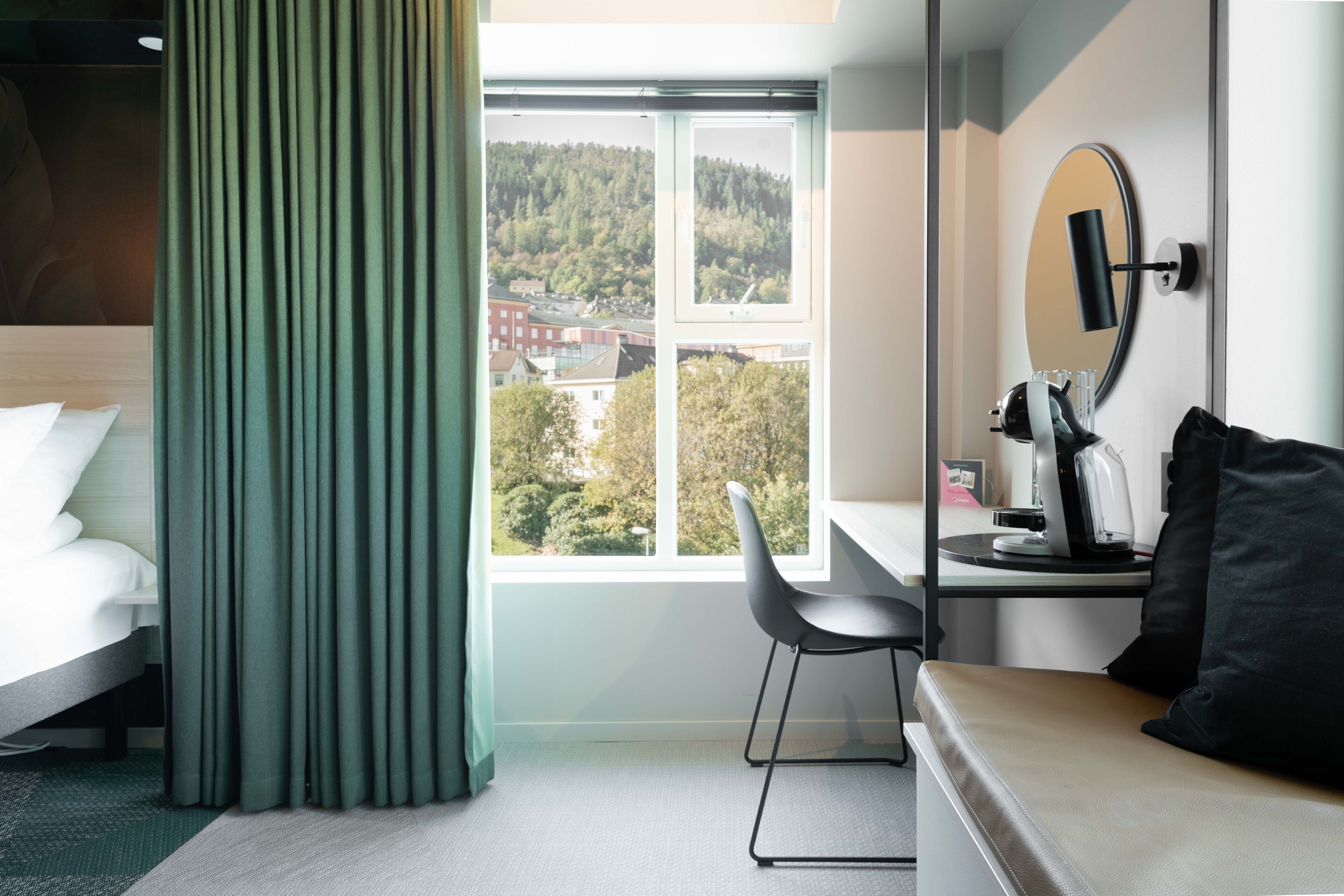 A hotel room with a bed, a desk with a chair, a mirror, and a view of a forested hill.