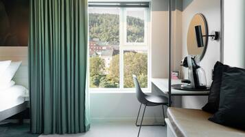 A hotel room with a bed, a desk with a chair, a mirror, and a view of a forested hill.