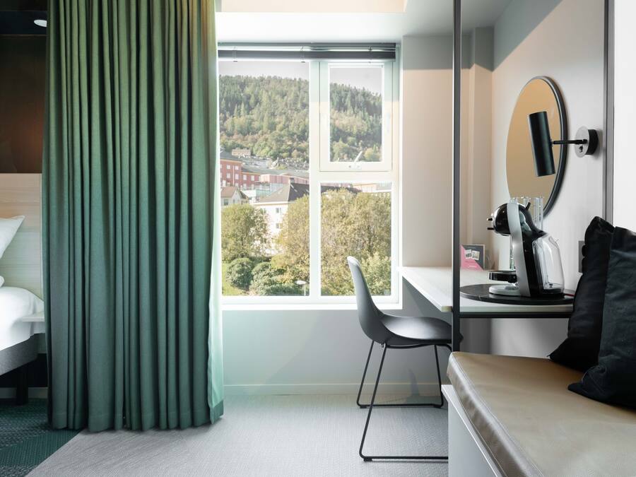 A hotel room with a bed, a desk with a chair, a mirror, and a view of a forested hill.