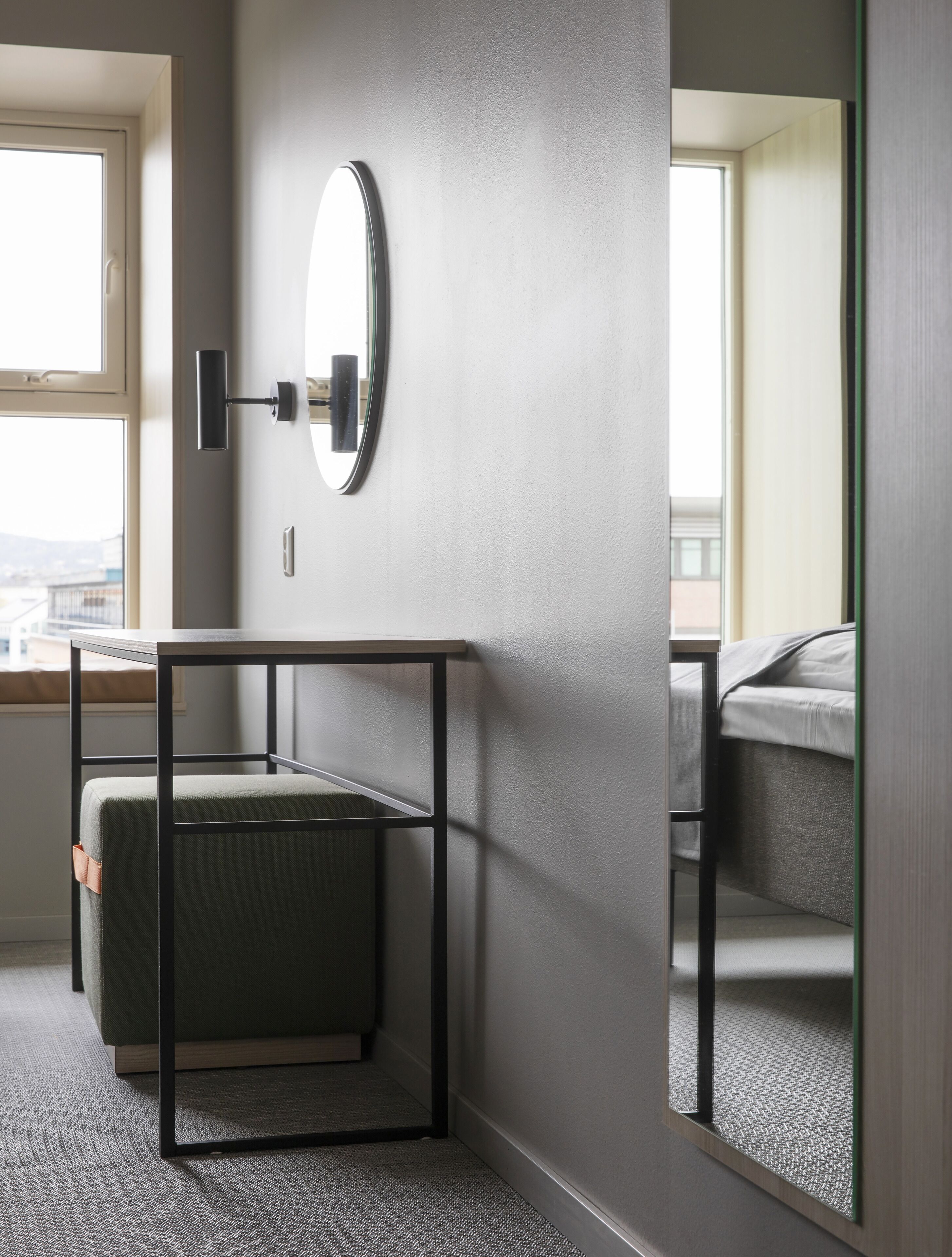 Une chambre d’hôtel moderne avec un mur gris, un miroir rond, une petite table avec un coussin vert et un lit recouvert d’une couverture grise.