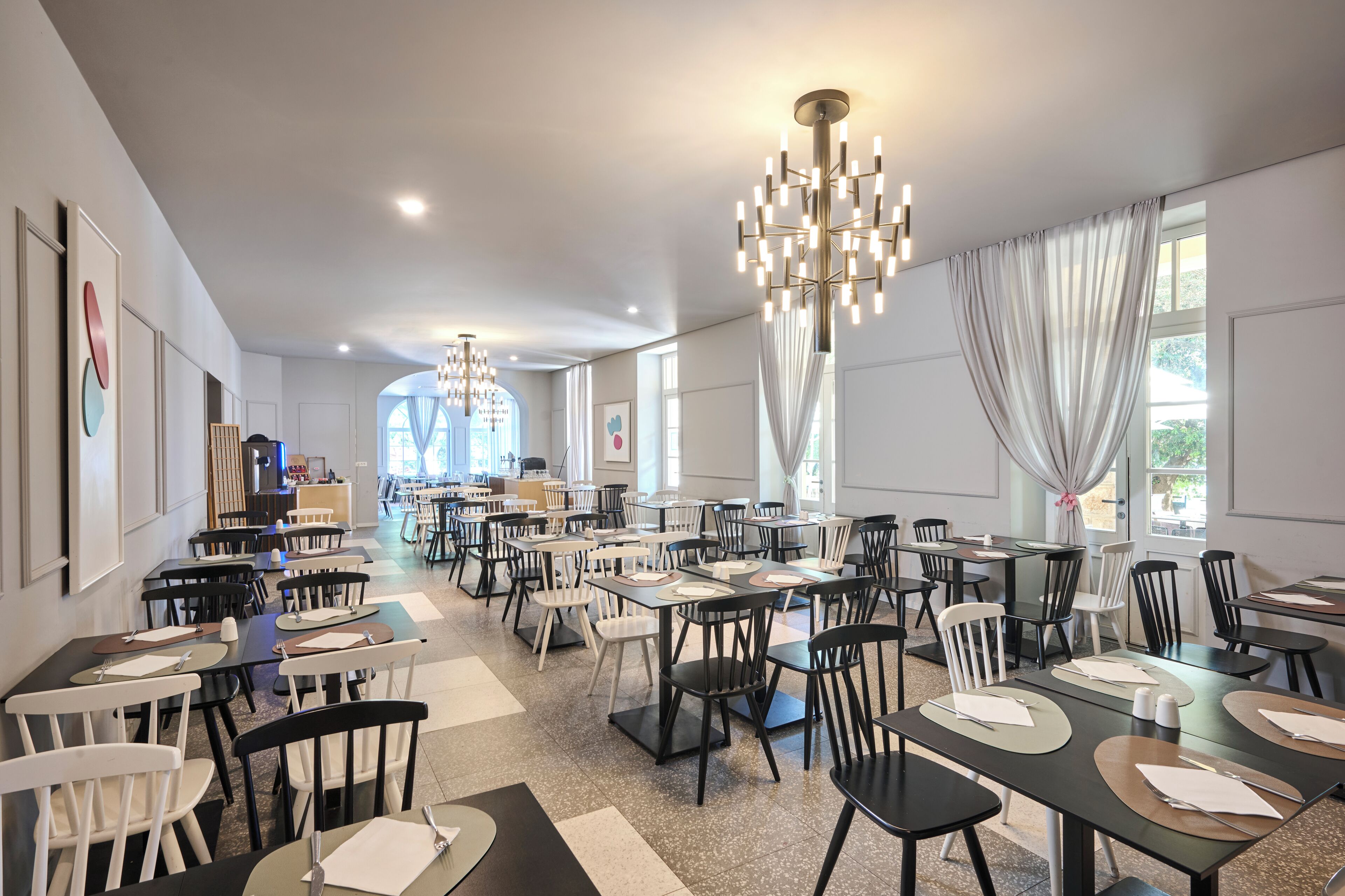 A spacious dining area with multiple tables and chairs, modern chandeliers, and large windows allowing natural light.
