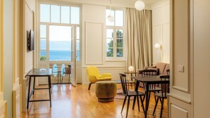 A spacious living area with a wooden floor, a yellow armchair, a round ottoman, a dining table with chairs, and a view of the sea through large windows.