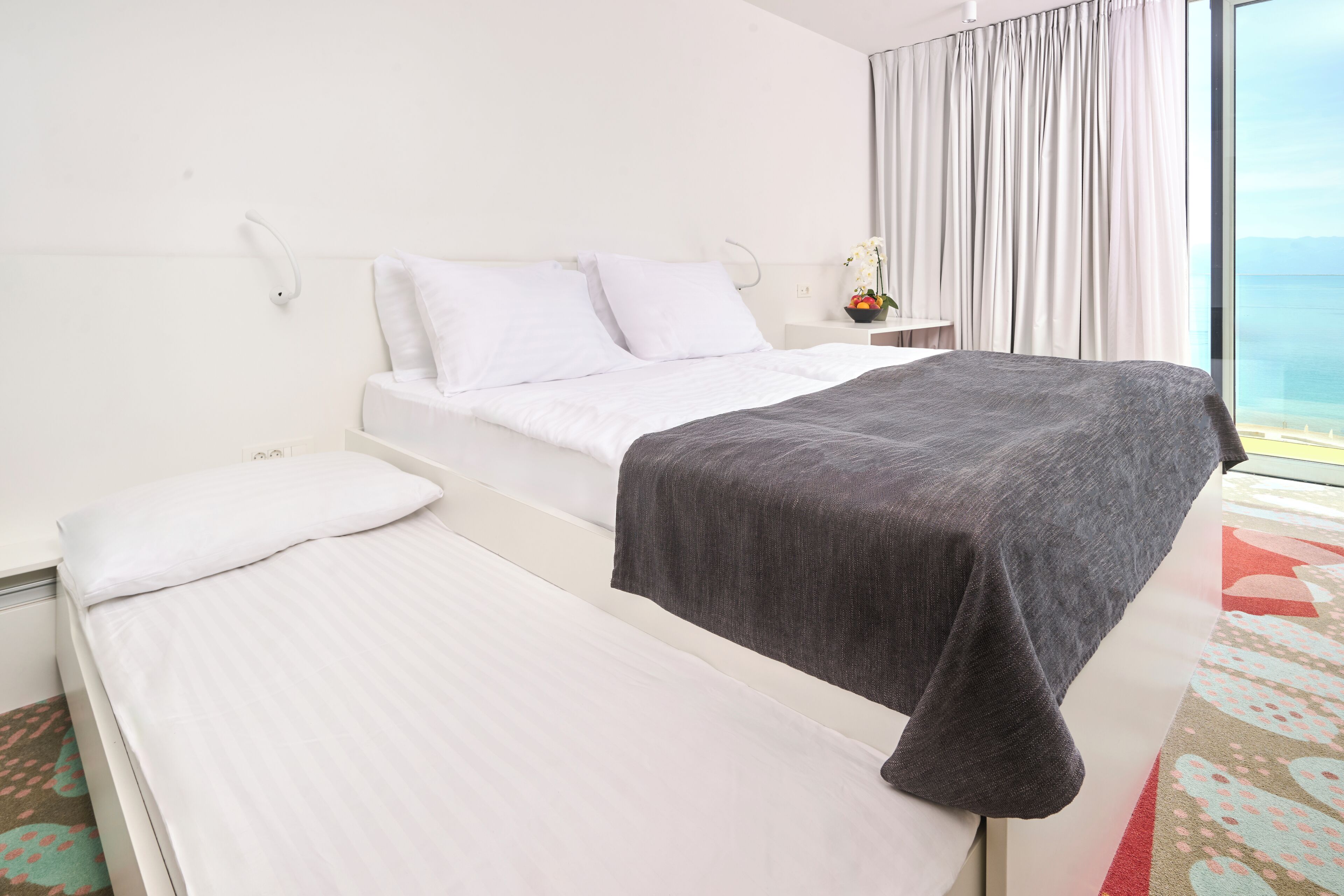 Camera d'albergo con un letto, un letto aggiuntivo o divano letto, un comodino con lampada, una finestra con tende e vista sul mare.