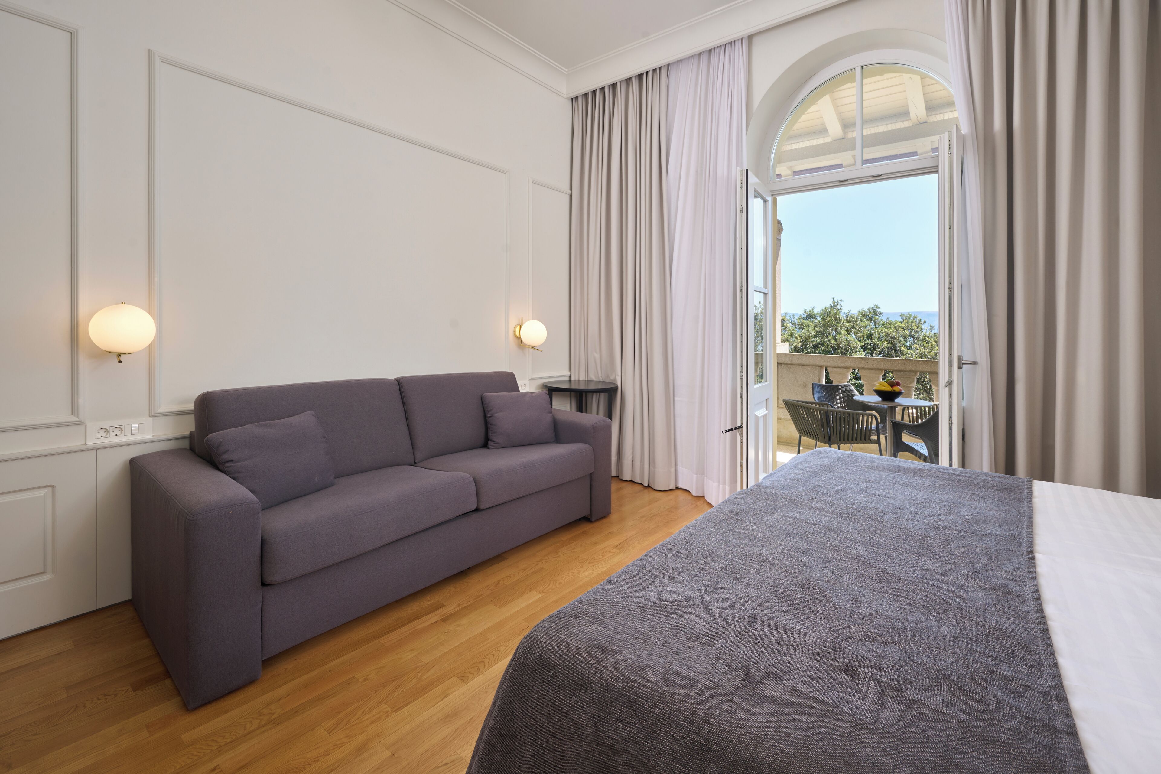 Una habitación de hotel moderna con un sofá gris, una cama grande y un balcón con mesa y sillas.