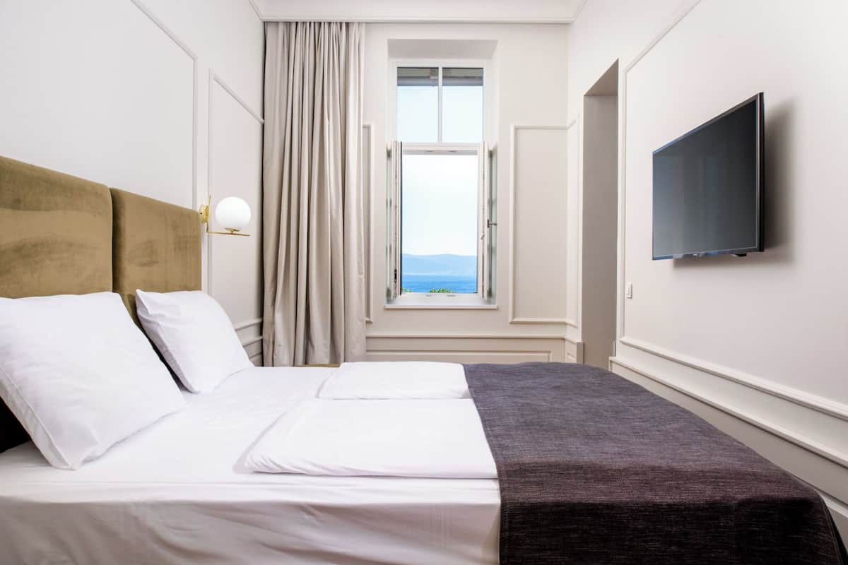 A hotel room with a bed, a TV on the wall, and a window with a view of the sea.