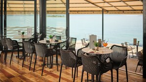 An indoor dining area with tables and chairs, large windows overlooking the sea, and a wooden floor.