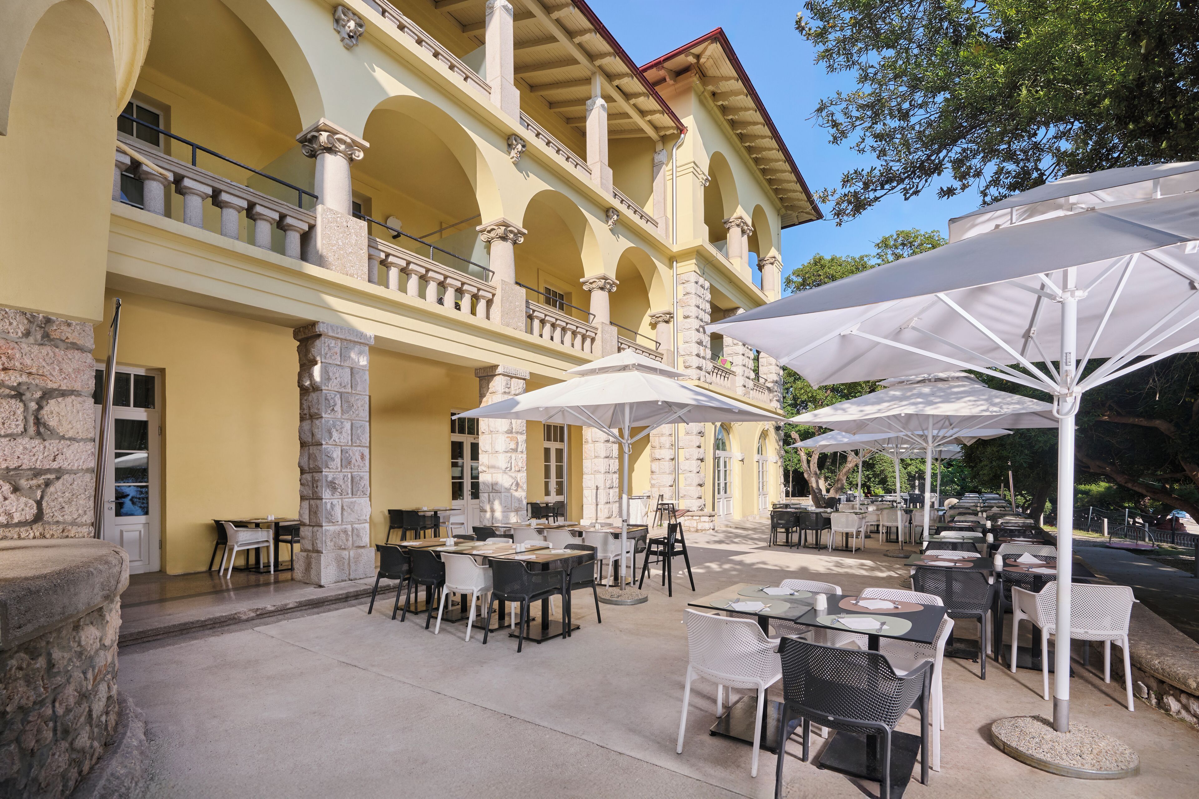 An outdoor patio with tables and chairs, large umbrellas, and a building with arches and columns.
