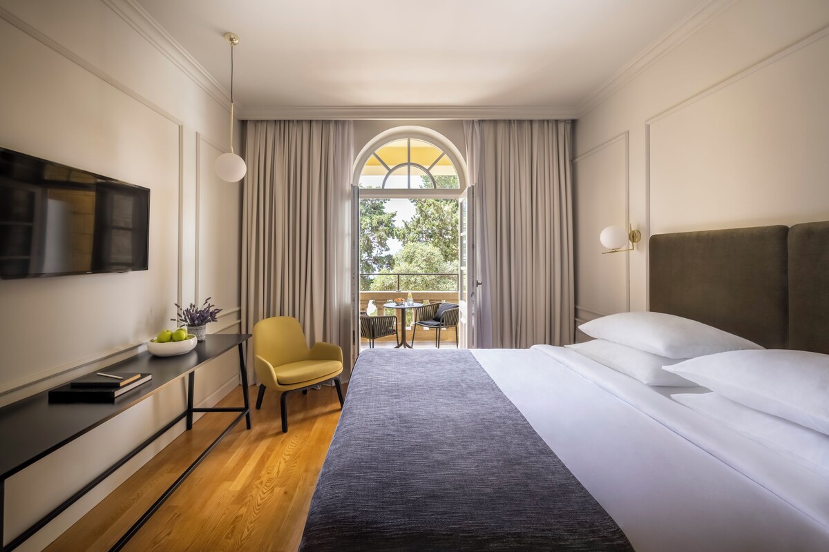 Habitación de hotel con cama, escritorio, silla, televisión y ventana con cortinas.