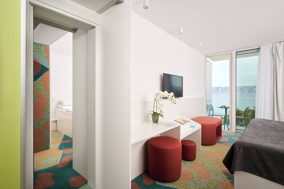 A modern hotel room with a bed, a desk, and a chair. There is a TV mounted on the wall and a window with a view of the ocean.