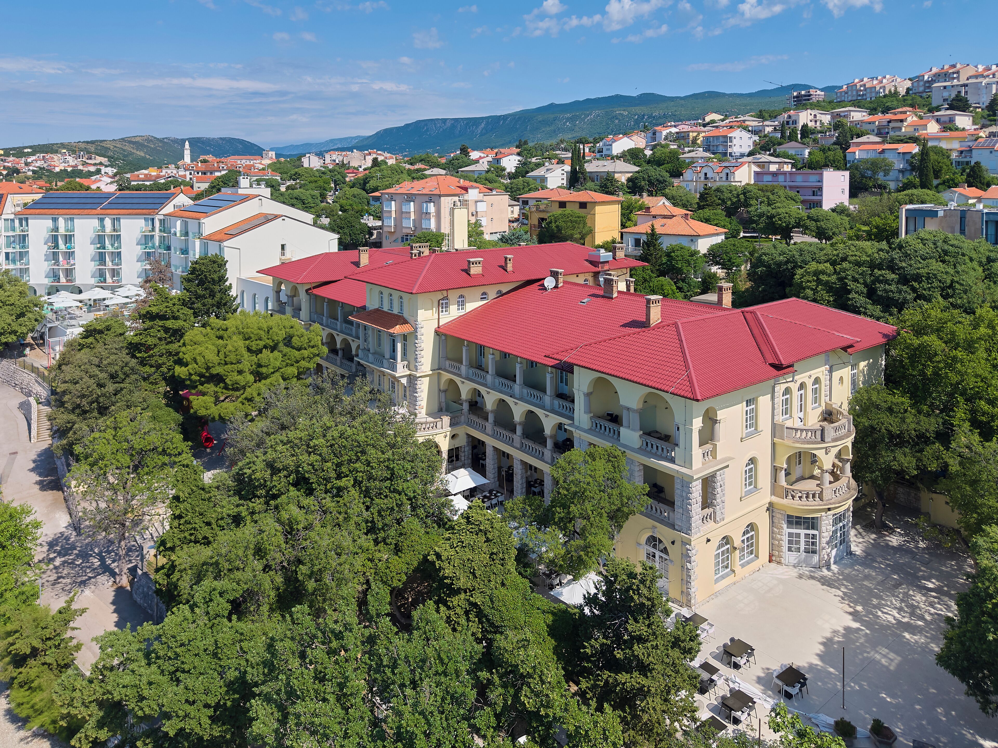 A large, multi-story building with red-tiled roofs, surrounded by lush greenery and other residential buildings.