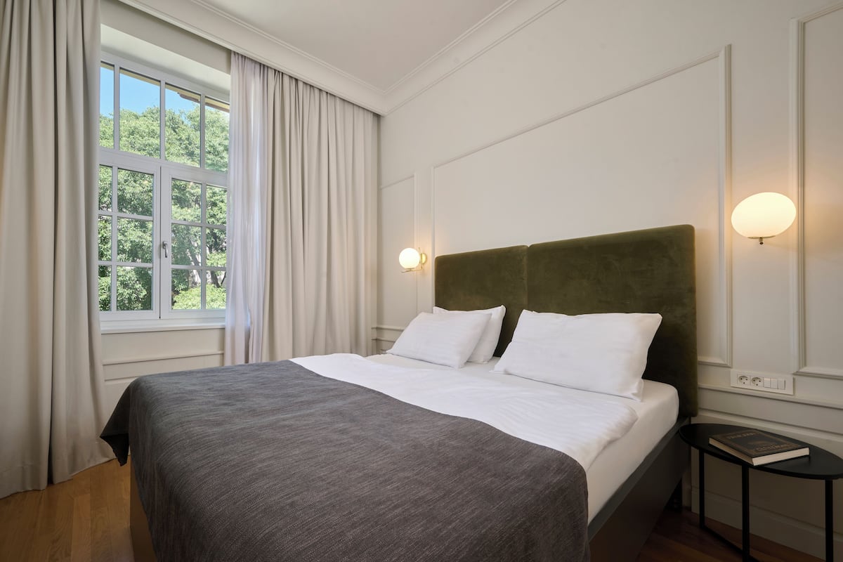A neatly made bed with white linens and a dark gray throw, a bedside table with a book, and a window with curtains.