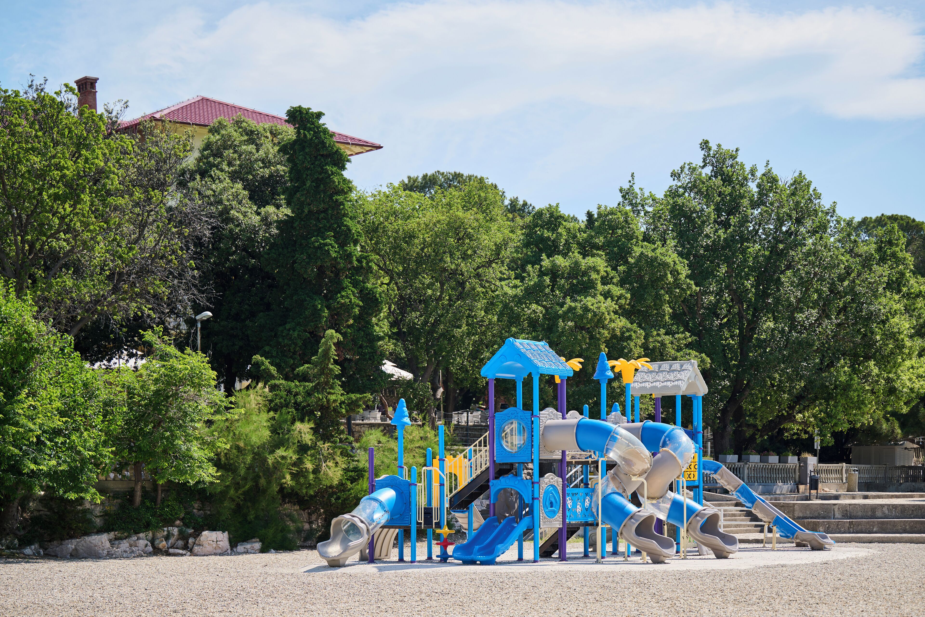 A playground with slides and climbing structures, surrounded by trees and a building with a red roof in the background.