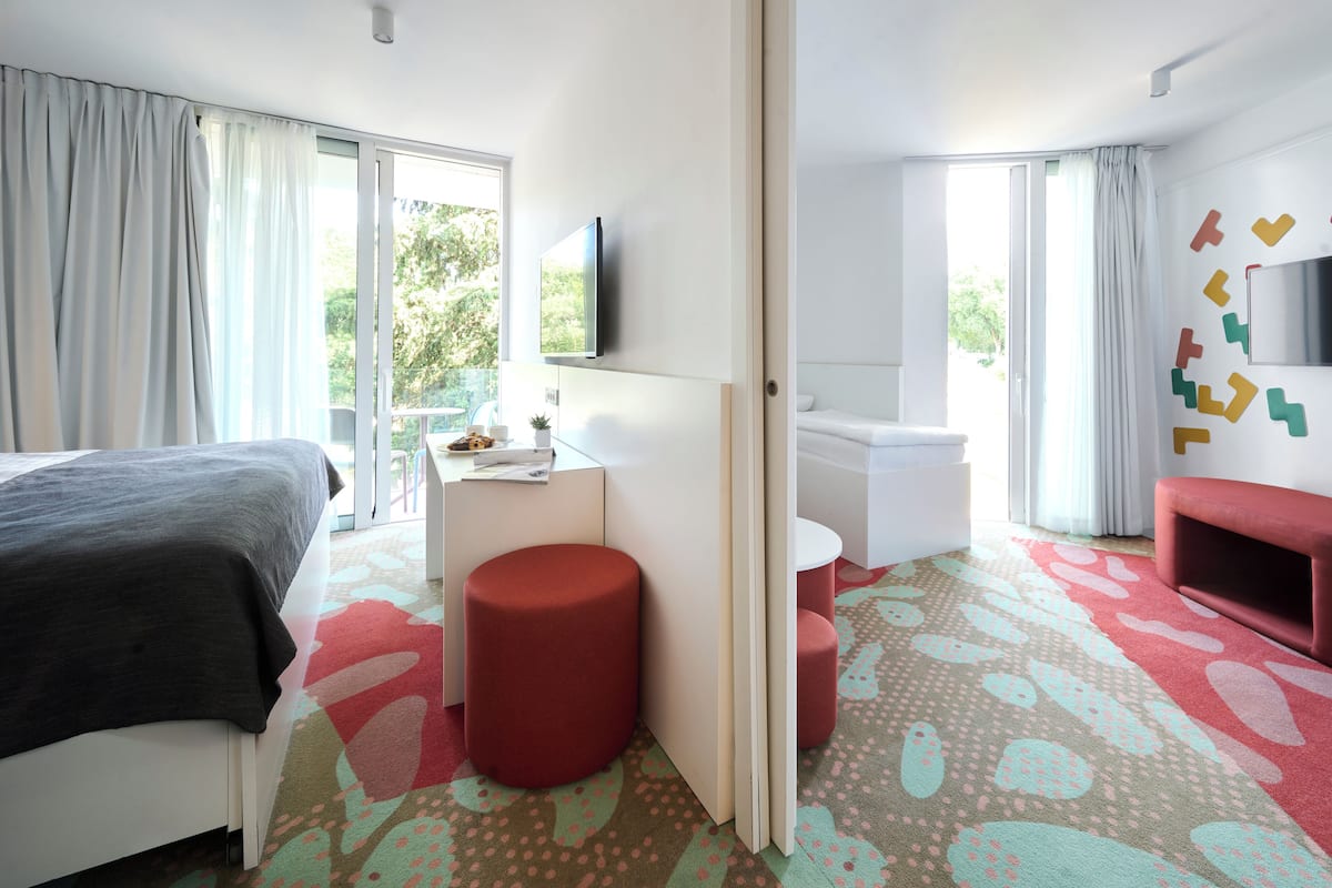 A hotel room with a bed, a desk, a red ottoman, a small round table, a red sofa, a TV mounted on the wall, and a patterned carpet.