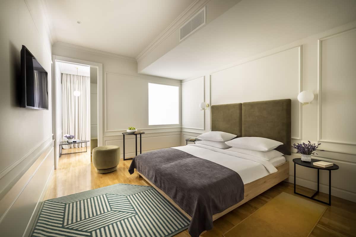 A modern bedroom with a large bed, bedside tables, a TV mounted on the wall, and a window providing natural light.